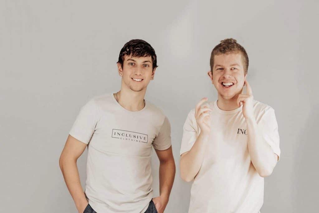 VIU student Brant Cartwright, left, stands beside his brother Riley with a white backdrop smiling in Inclusive Clothing T shirts