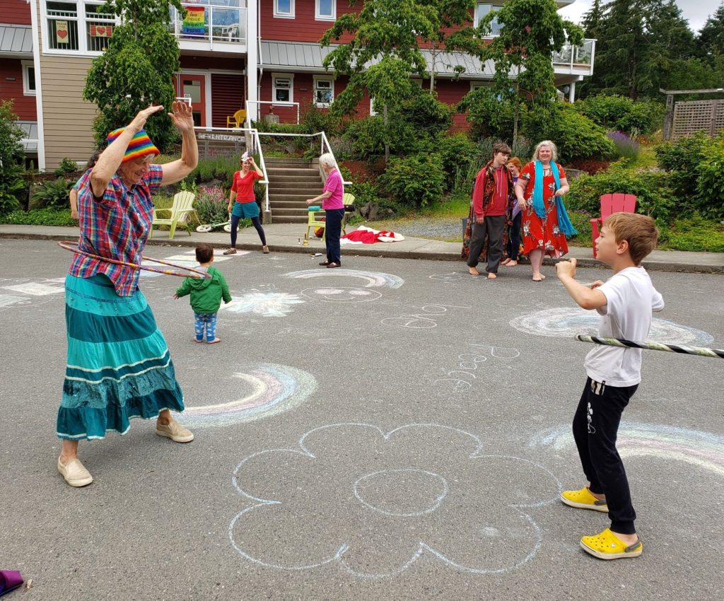 An older person and a young person stand outside facing eachother playing with hulahoops at Nanaimo's only cohousing complex, Pacific Gardens.