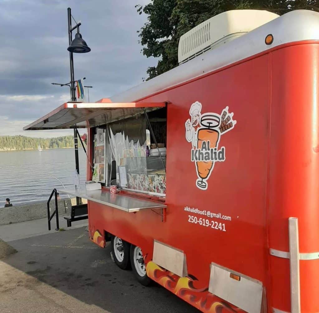 A red Shawarma food truck with the words Al Khalid is parked at a beach.