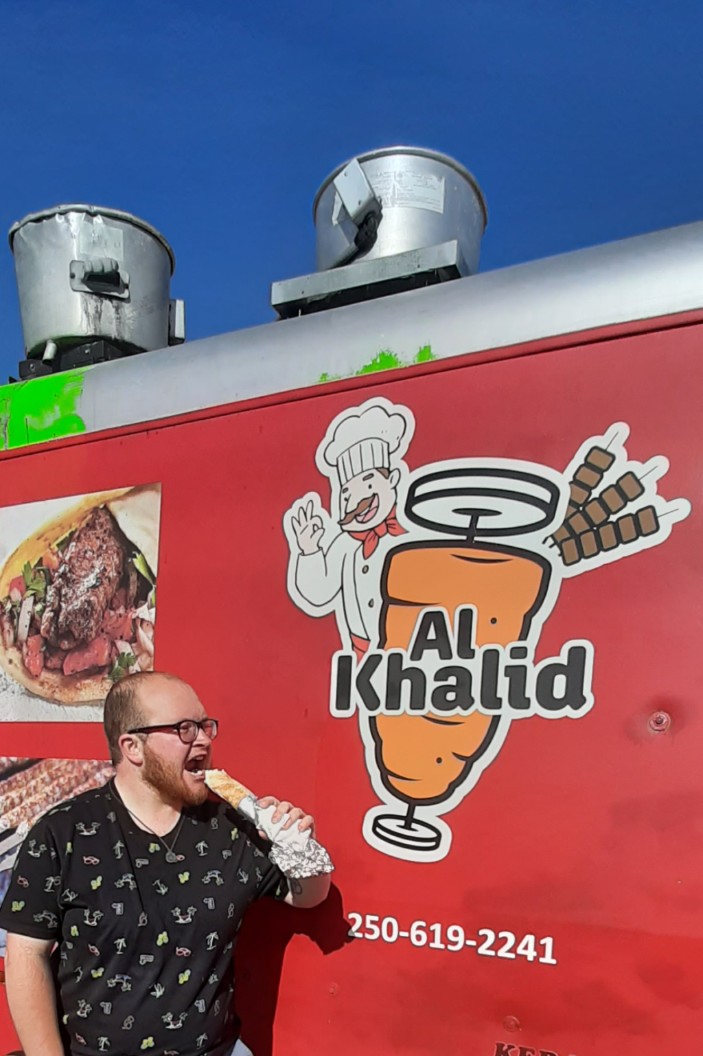 a man stands in front of a red food truck, over exageratingly taking a bite out of a pita wrap sandwich.