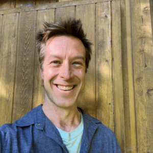 A selfie of Andrew Rushmere smiling, from the shoulders up. He is standing in front of a wood panel wall and wears a dark blue collared shirt with a light blue shirt underneath.