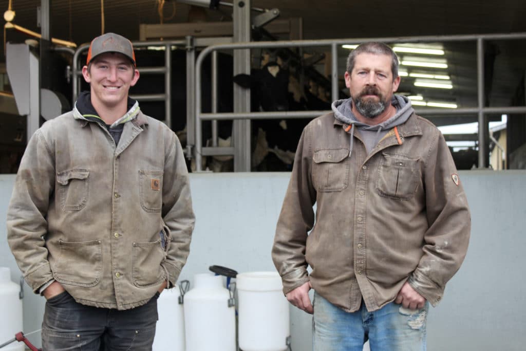 Dairy farmers Ian and Jack porter stand in front of a barn with cows in the background. The elder Ian is on the right and Jack is on the left. They're both wearing brown jackets and have their hands in their pockets. Ian has a beard and neutral expression, Jake is smiling and is wearing a brown ball cap.