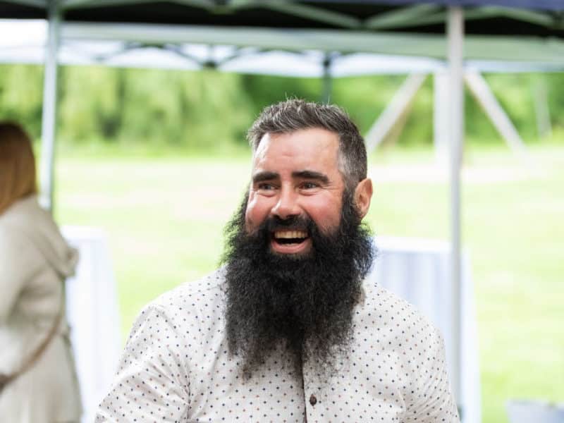 Bill Keserich Junior is pictured at the centre of this photo, from chest-up. He is wearing a white, short-sleeve, collared button-up that has many small, coloured diamond shapes printed on it. His beard is shoulder-length and black and he is smiling in the photo. Behind him is a green field.