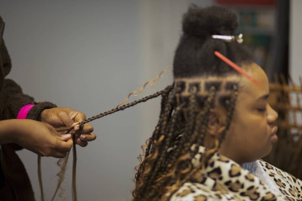 A womans hands are shown braiding curly strands of human hair into long braids, the clients hair is long so the braids extend down her back.