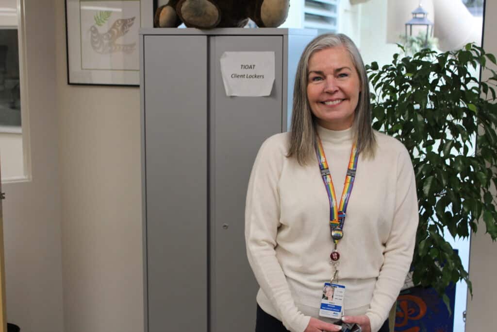 Jessica Huston, who oversees the wellness centre, poses for a photo. She is pictured from waist-up, smiling at the camera. She wears a long-sleeve shirt and a name tag hangs around her neck. Behind her is a metal cabinet to the left and a plant to the right.