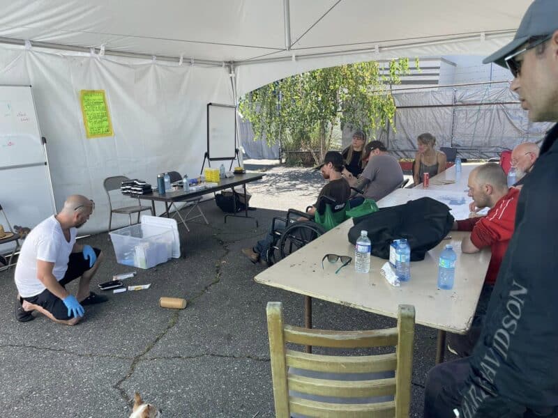 Members of NANDU, a peer run drug consumption group, sit in chairs watching a nurse demonstrate how to administer Naloxone.