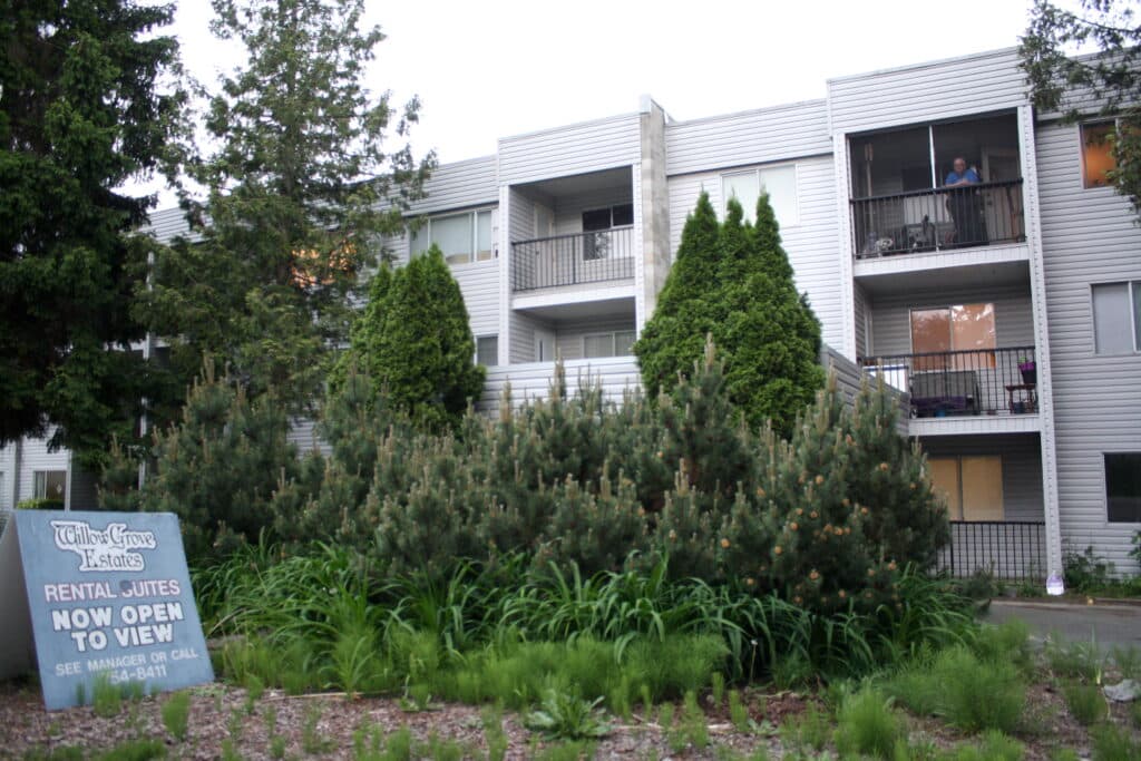 An older rental building with four storeys and balconies is nestled in trees. A sign reads Willow Grove Estate rental suites now open to view.