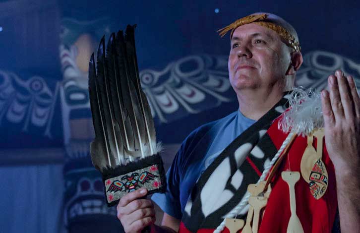 Daniel Elliott of Raven Sky Productions holds eagle feathers against a backdrop of eagles. He is wearing a cedar headband and blanket.