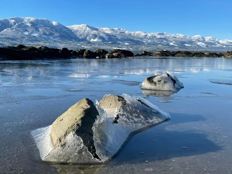 Ice breaks over rocks in the ocean with snowy mountains in the distance.