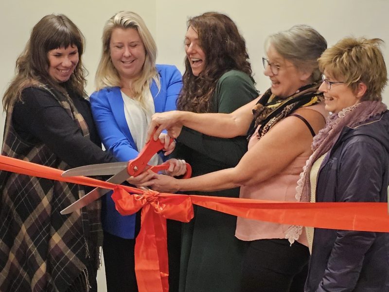 Five women gather in front of a large red ribbon, each holding a large pair of red scissors as they cut the ribbon. They all carry expressions of excitement and happiness on their faces.