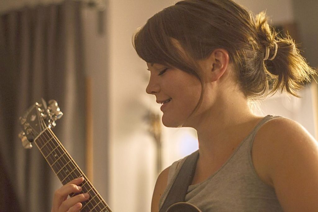 A young woman with brown hair and a grey tank top plays guitar and sings