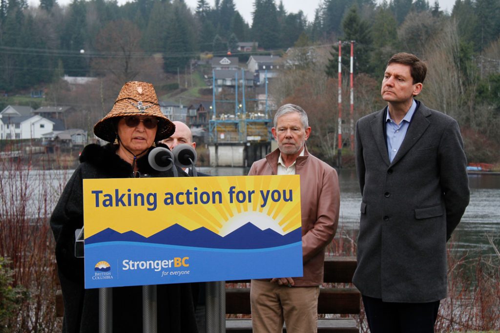 A woman in a women Coast Salish hat stands at a podium and three men stand behind