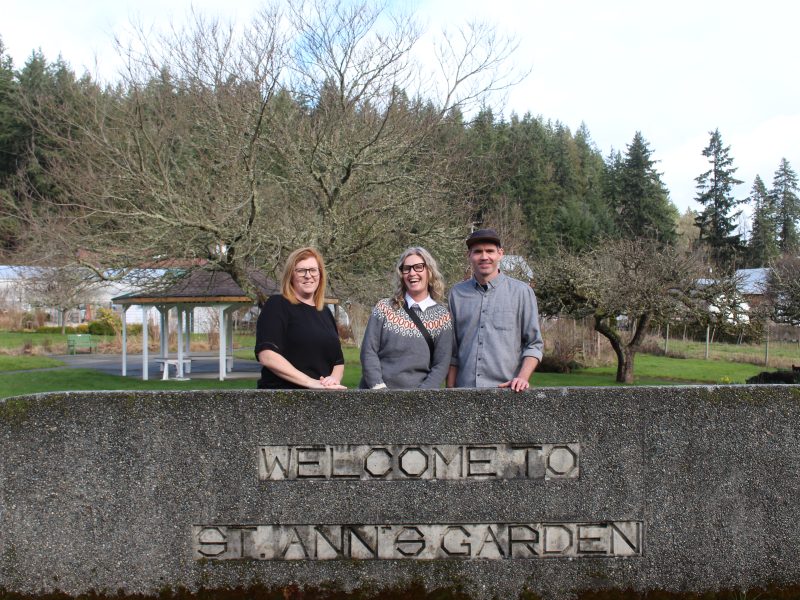Leah Boisvert, Stephanie Kok and Conrad Murphy stand behind a cement half-wall that has St. Ann's Garden Club engraved in it. Behind them is a field with walking paths and apple trees.