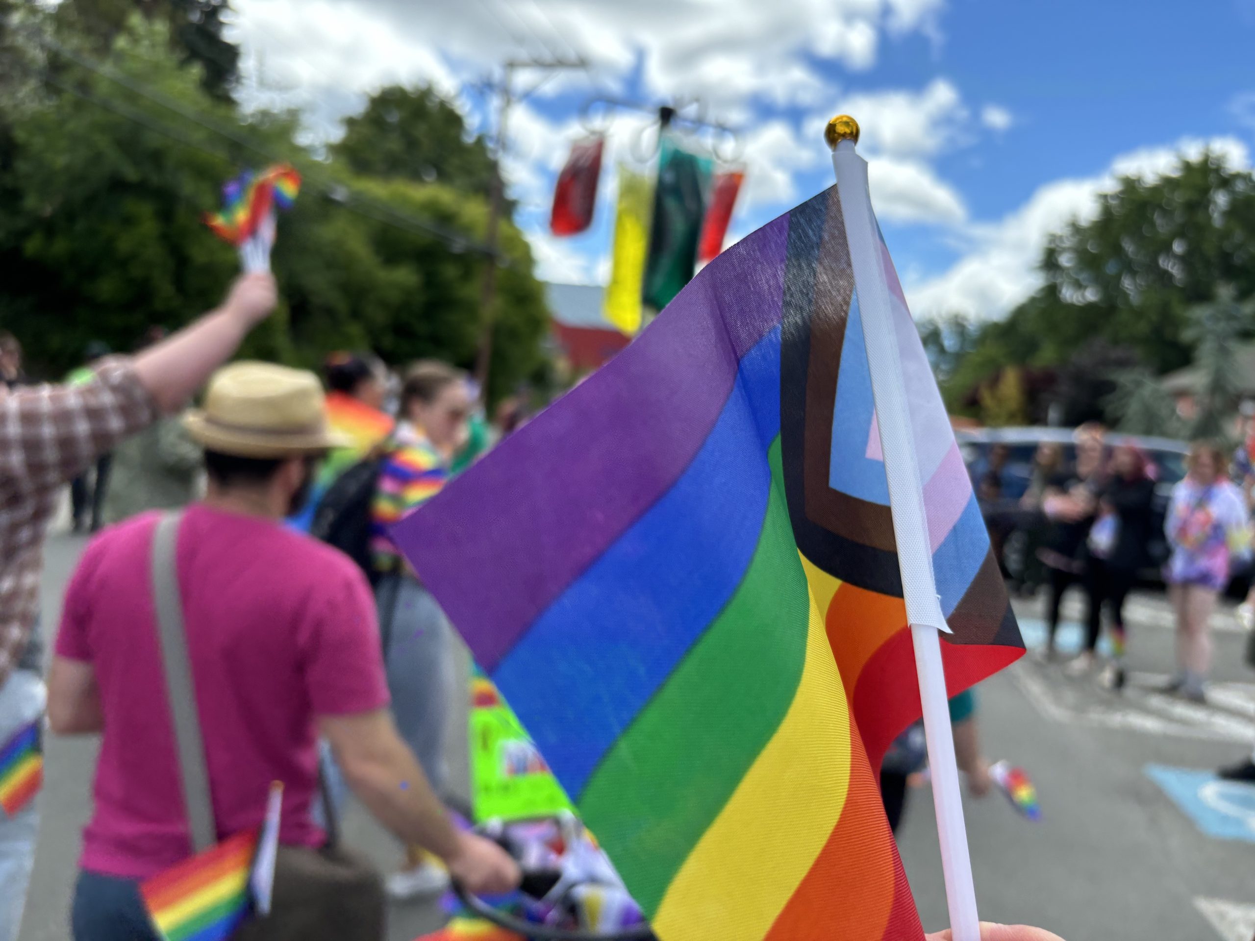 A rainbow Pride Progress flag waves with Pride parade goers in the background