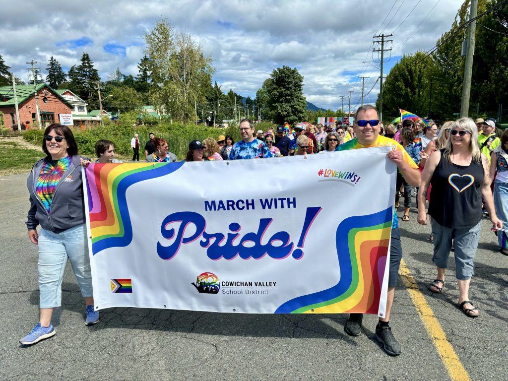 Two people stand on either side of a Cowichan Valley School District banner as they walk through the Pride parade, followed by a large group of people.