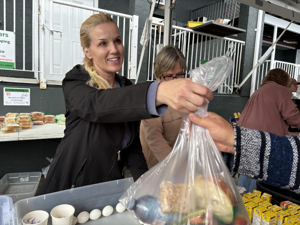 Simone Conner hands a plastic bag containing vegetables, a sandwich, snacks, and water to a guest at Basket Society's Sunday Sandwiches program